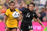 A Matildas footballer and a New Zealand player jostle each other for position as the ball bounces in front of them.
