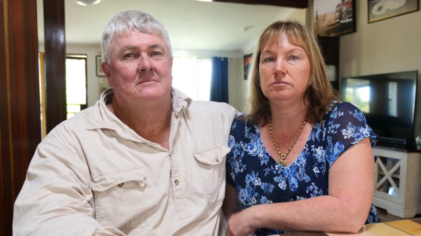 a man and woman with arm in arm at a dining table inside their home, looking at the camera with serious expressions