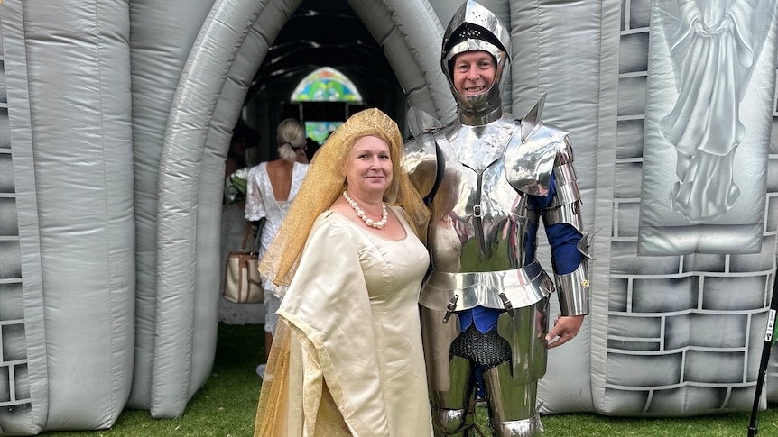 woman in gold dress and headress arm in arm with man in knight's armour in front of inflatable church