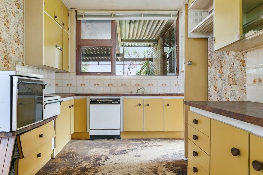 A dated kitchen with damaged yellow cupboards and floral wallpaper.