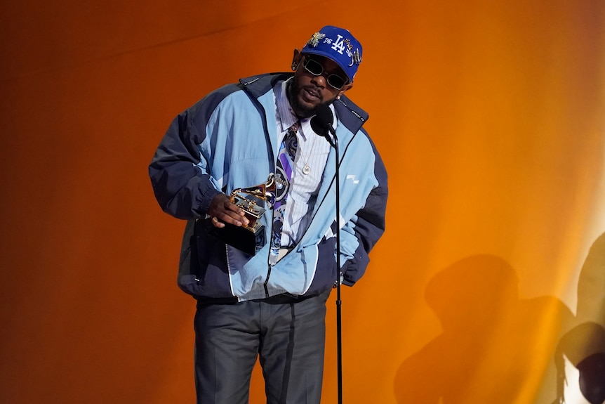 A young black man in a blue jacket and cap leans into the microphone while holding a trophy onstage at the Grammys.