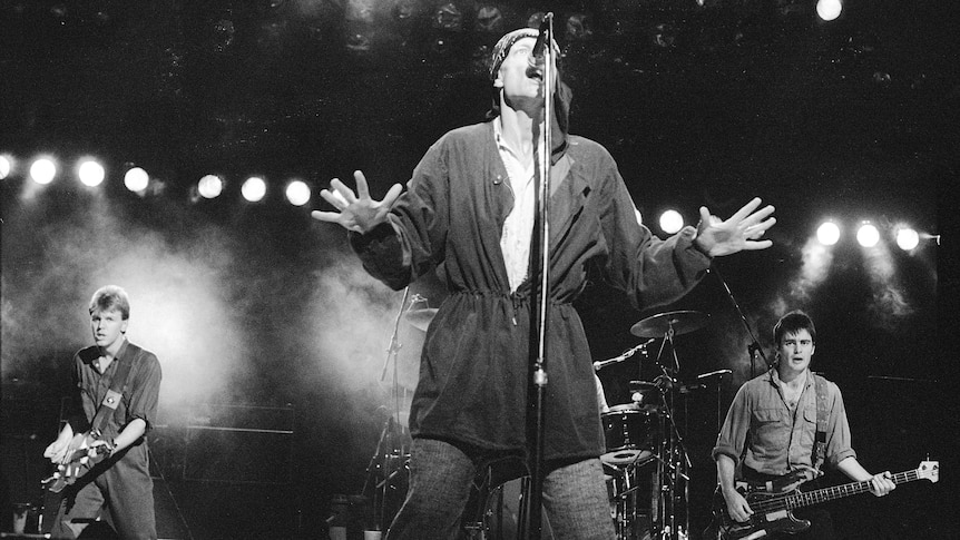 Black and white photo of Peter Garrett wearing headscarf singing into microphone. The band plays behind him.