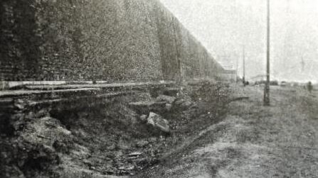 A black and white photo of a ditch between poles and a wall.
