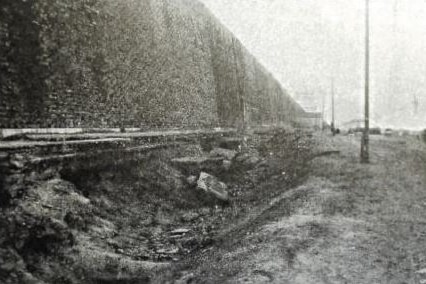 A black and white photo of a ditch between poles and a wall.