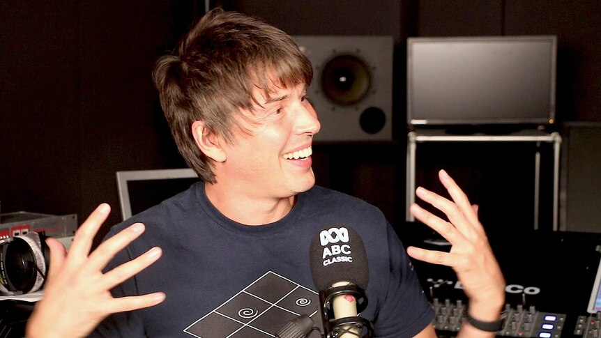 Brian Cox looks slightly to the side with an animated expression and both hands gesturing. There is a microphone in front of him