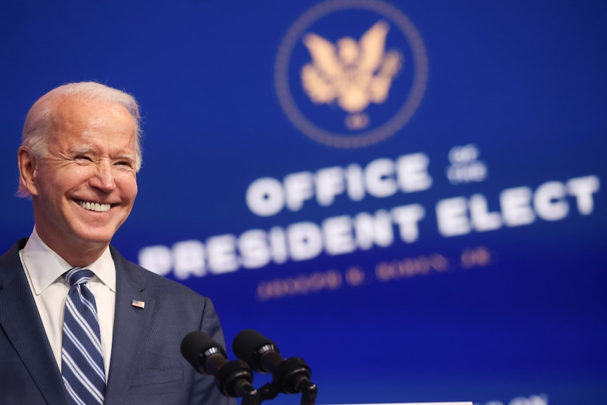 Joe Biden smiling as he stands in front of a "Office of the President Elect" sign in Delaware.