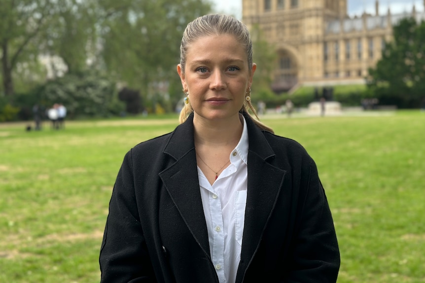 Blonde woman wearing light blue shirt and black coat standing in front of Britain's House of Parliament