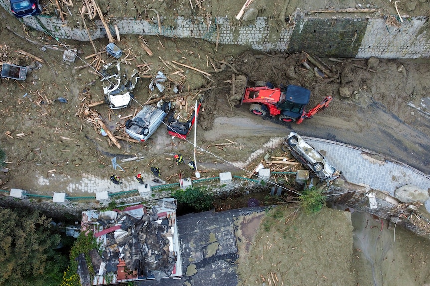 An overhead shot of a small bulldozer pushing mud and debrus out of a flooded road, where trees and overturned cars lay. 