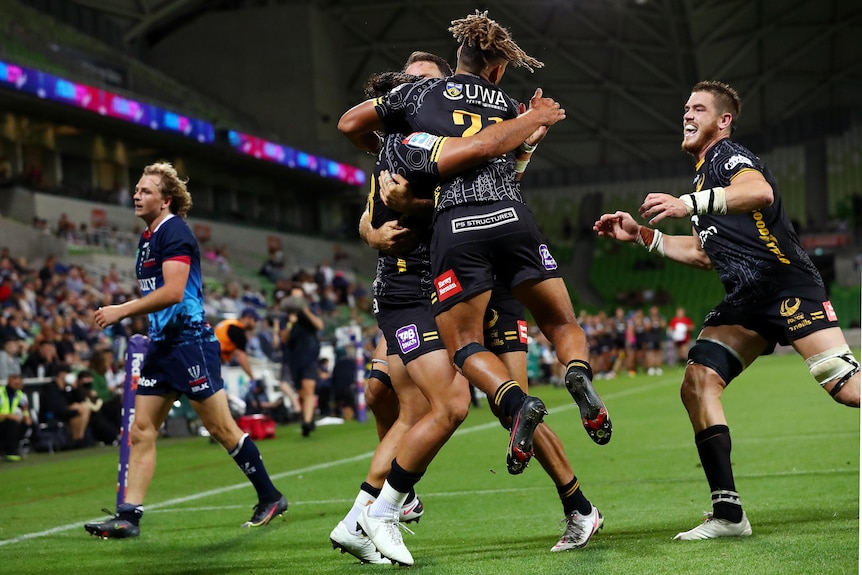 A group of happy Super Rugby teammates jump into each others' arms to celebrate a try as a dejected defender stands to one side.
