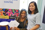 Two women sit together holding letters near a colourful multicultural centre sign