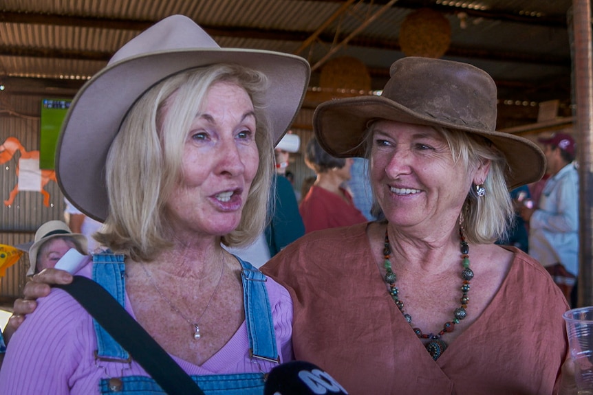 Lady in pink shirt and beige hat chats into a microphone while standing next to her female friend