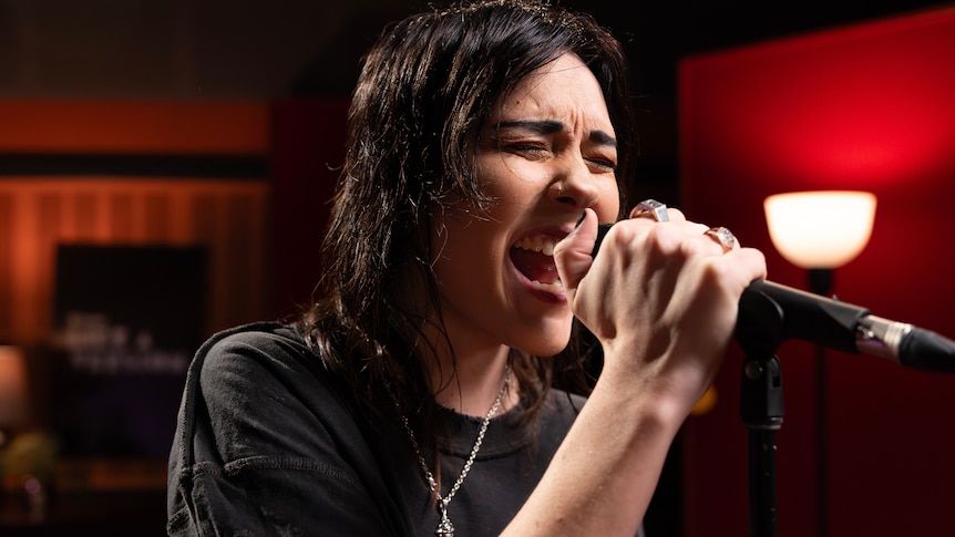 Person holding and singing into a microphone in a studio. They are wearing a black t-shirt