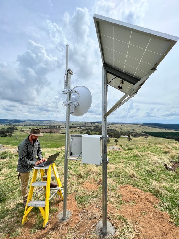 A man with a laptop stands near some apparatus on a farm.