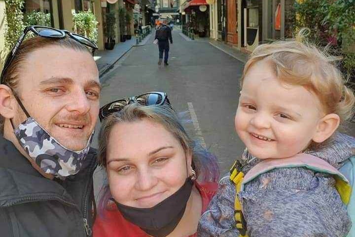 A man, woman and young child smile closely to camera with city alleyway behind them