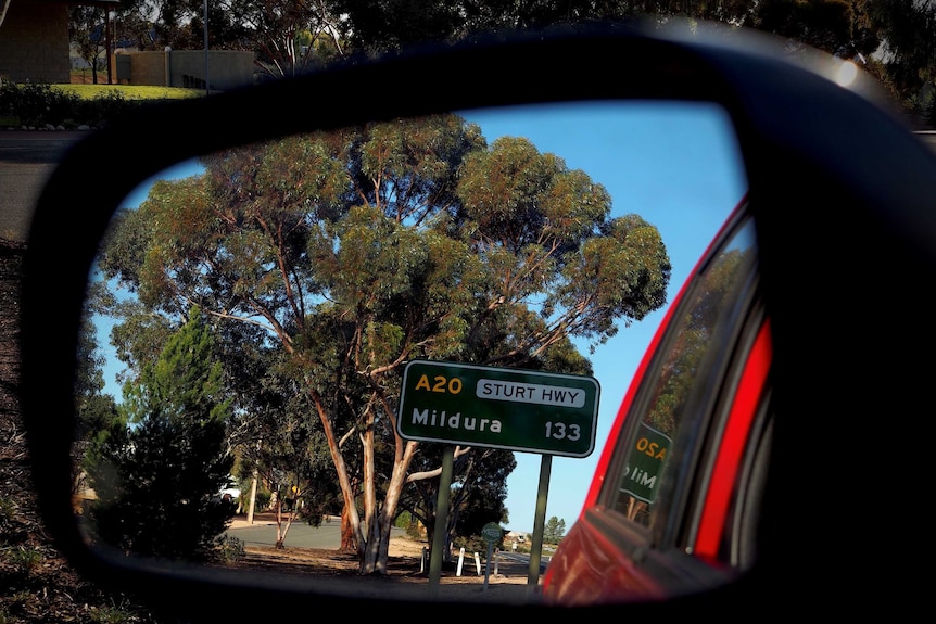 A green road sign reading Sturt Hwy, Mildura, 133 kilometres is captured in the side mirror of a red car.