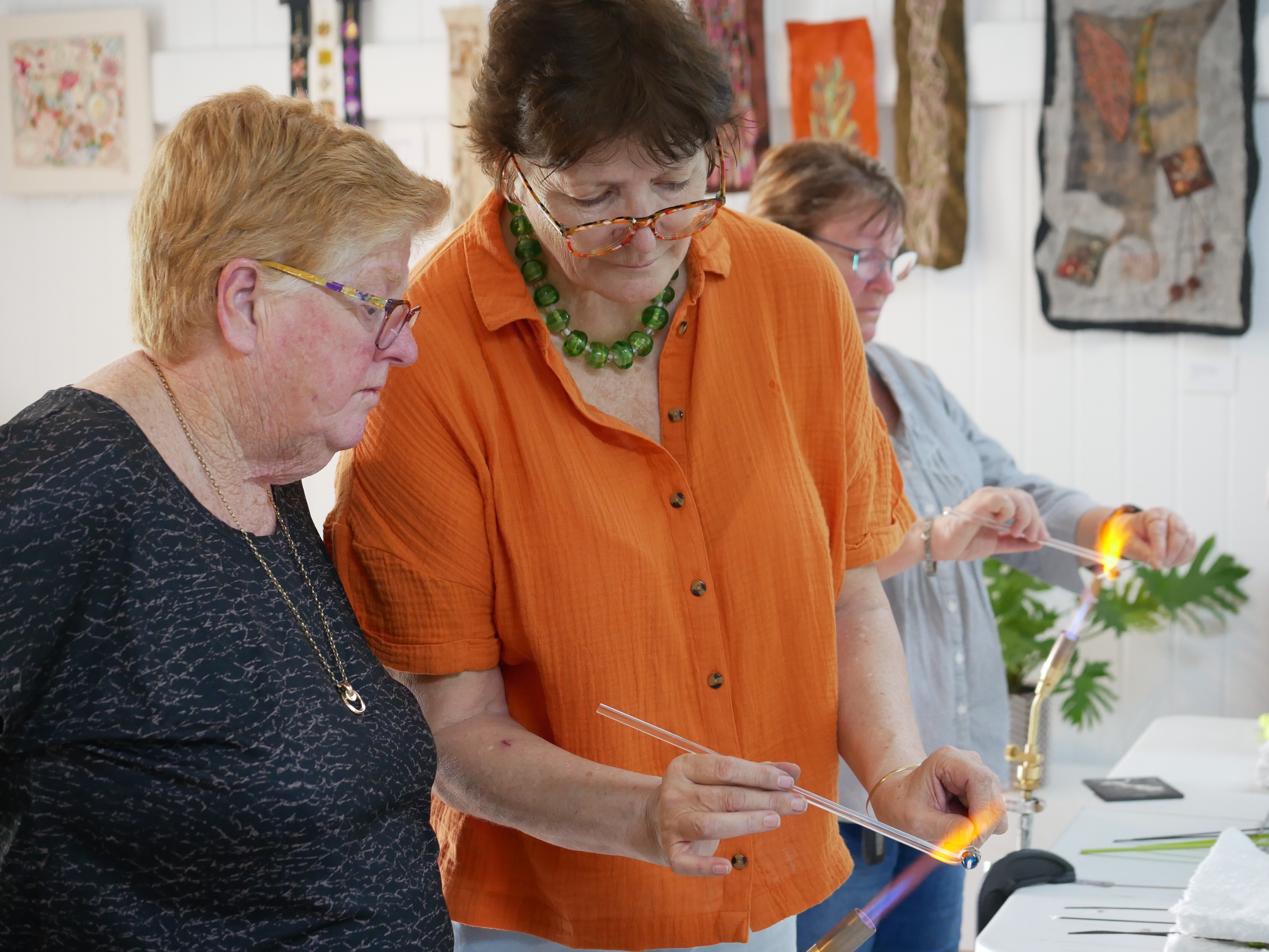 A woman in an orange shirt shows another woman how to mould glass over a flame