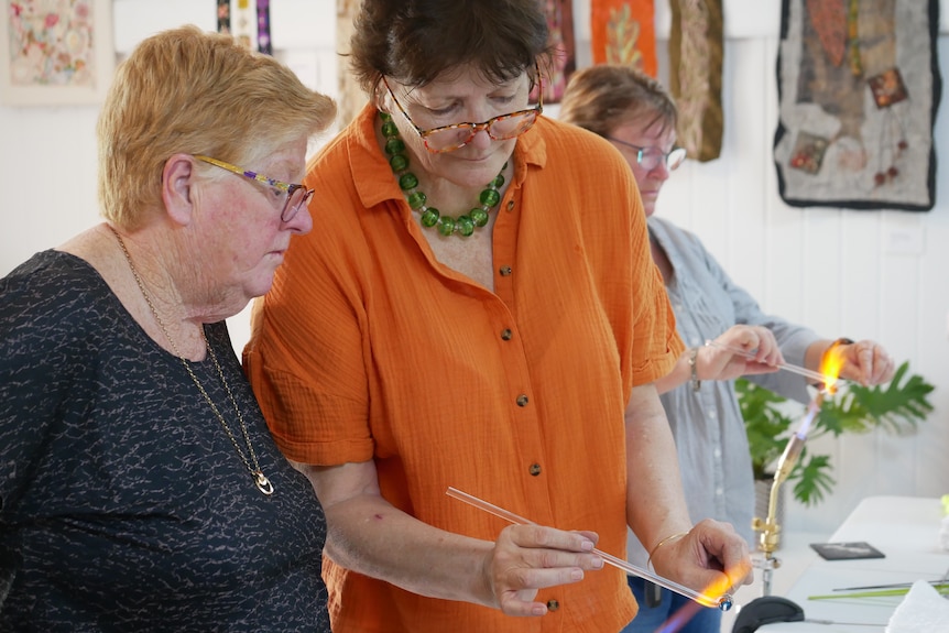 A woman in an orange shirt shows another woman how to mould glass over a flame