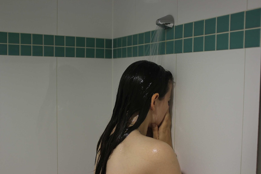 A young woman facing the bathroom tiles while standing underneath the shower.