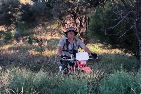 A white man in his fifties wearing a bucket hat, t-shirt and shorts on a red trail bike in high grass on a rural proeprty.