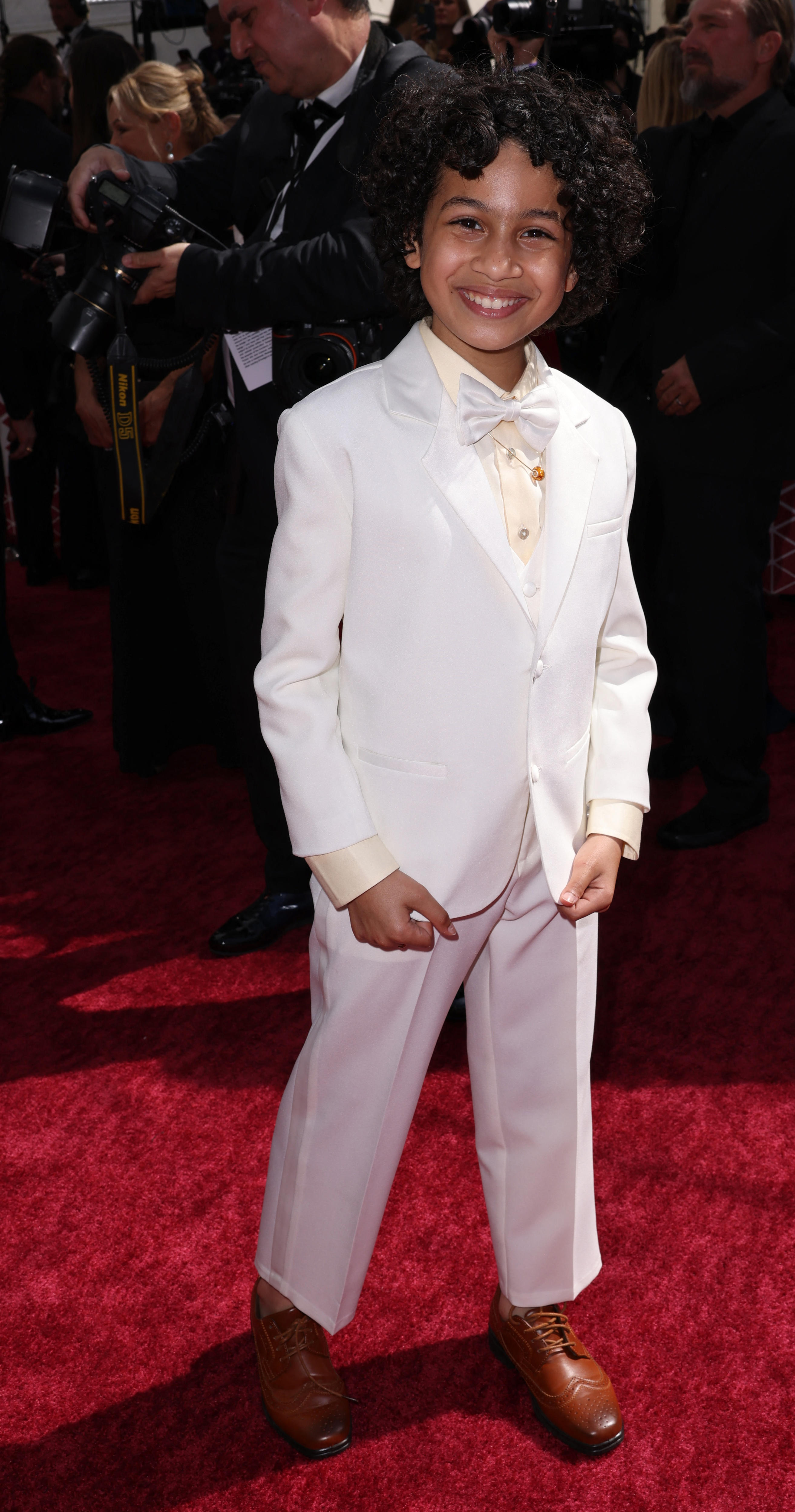 Child actor Ravi Cabot-Conyers smiles on the Oscars red carpet wearing a white suit with bow tie.