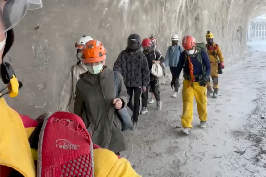 A group of people and rescue workers wearing helmets walk along a grey tunnel.
