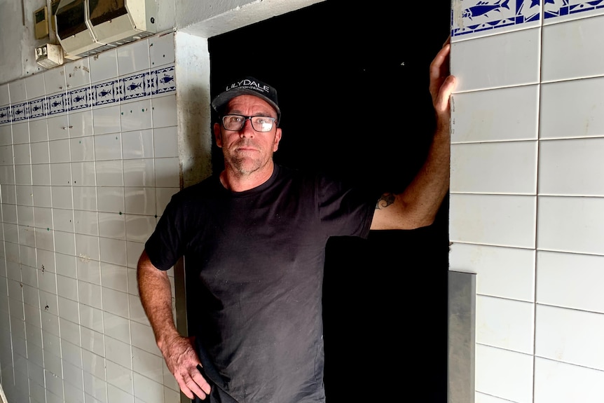 A man in a black t-shirt stands in a doorway surrounded by white tiled walls.
