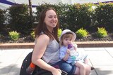 A woman in a wheelchair with her young daughter on lap on a footpath, shrubs and building in background.
