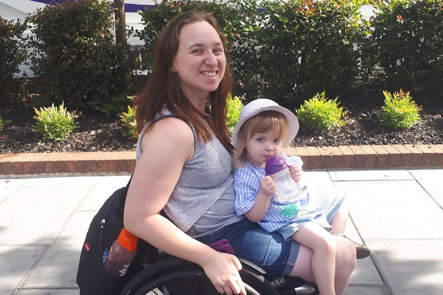 A woman in a wheelchair with her young daughter on lap on a footpath, shrubs and building in background