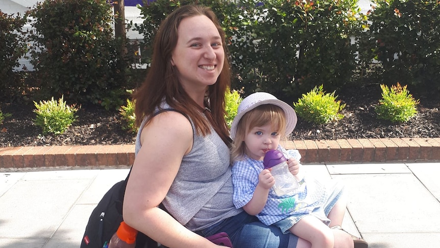 A woman in a wheelchair with her young daughter on lap on a footpath, shrubs and building in background.