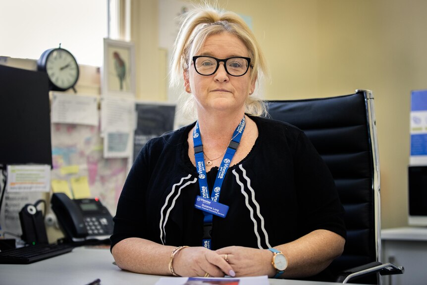 A woman in glasses and a blue lanyard sits at a desk in a cluttered office.