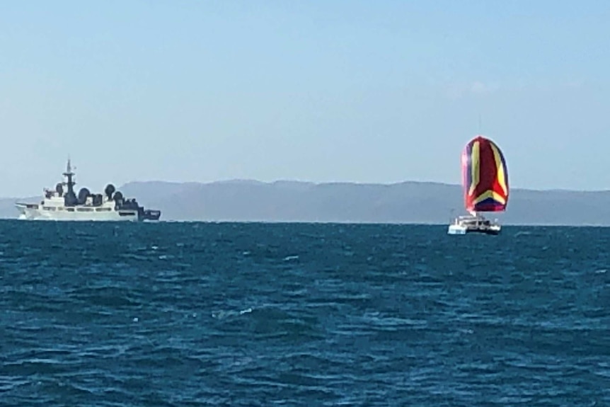 A ship is seen in the far left corner of the image with a boat to the right with a billowing, red, purple and yellow sail.