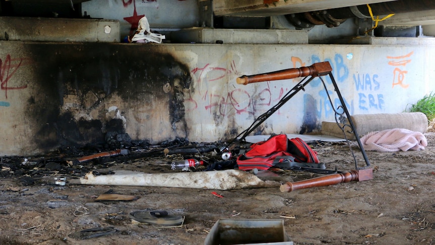 A bed frame sits under a bridge, half burned by people who are homeless.