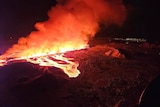Bright orange liquid lava flowing down a hill with smoke risign above late at night