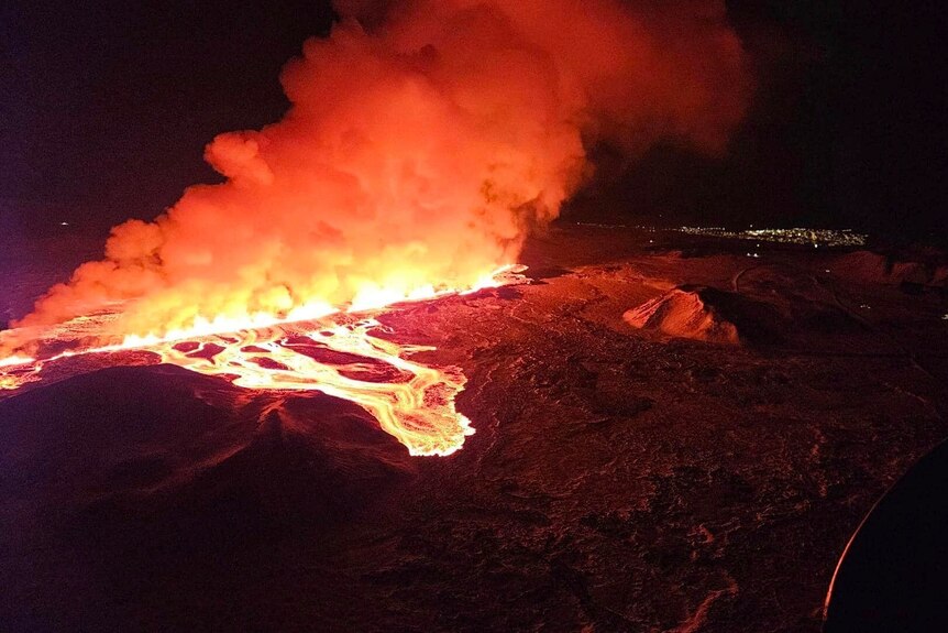 Bright orange liquid lava flowing down a hill with smoke risign above late at night