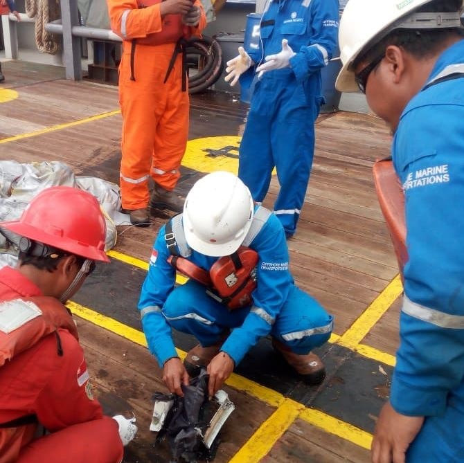 Men in helmets and safety clothing on a boat look at items on the deck.