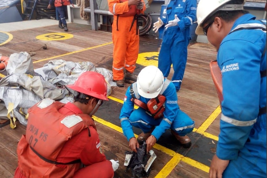 Men in helmets and safety clothing on a boat look at items on the deck.