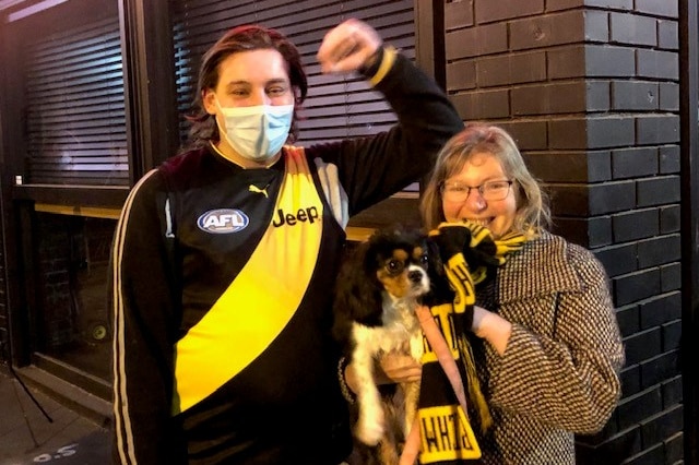 A young man pumps his fist in the air while a woman in a black and yellow scarf grins and holds a small dog.