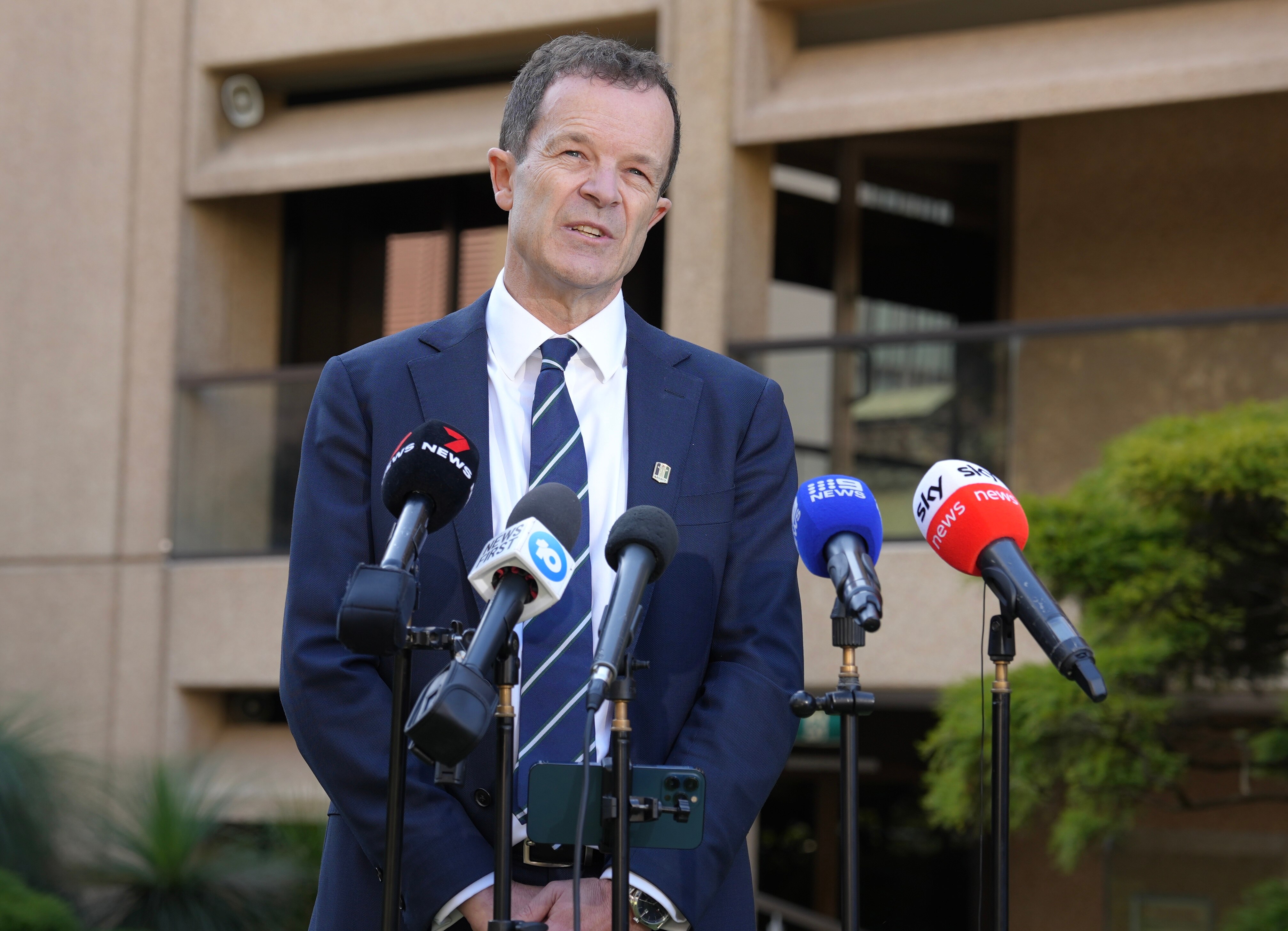 A middle-aged man in a suit speaks to the media outside a building.