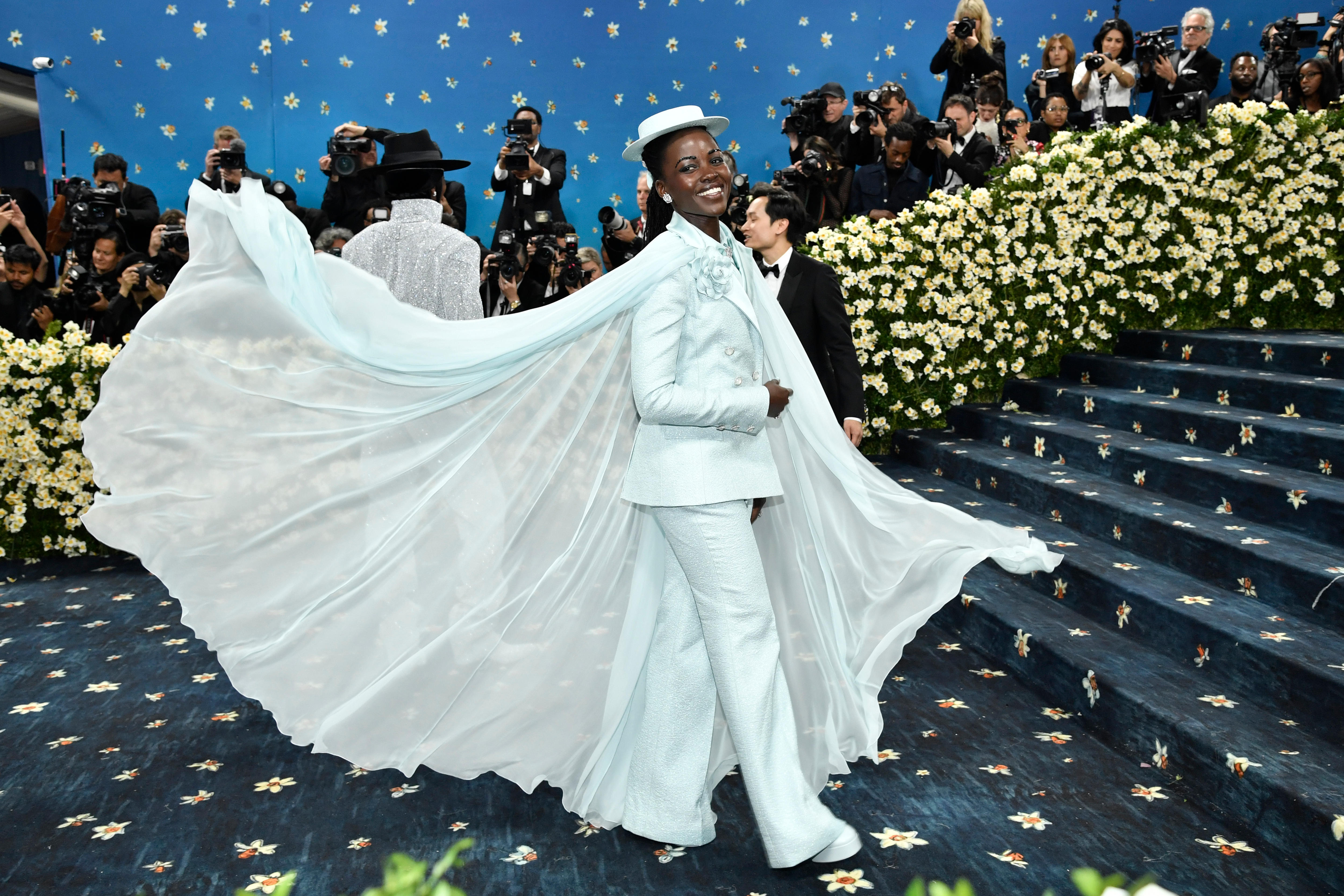 Lupita Nyong'o wearing a pale blue suit with a matching boater hat and a soft sheer cape