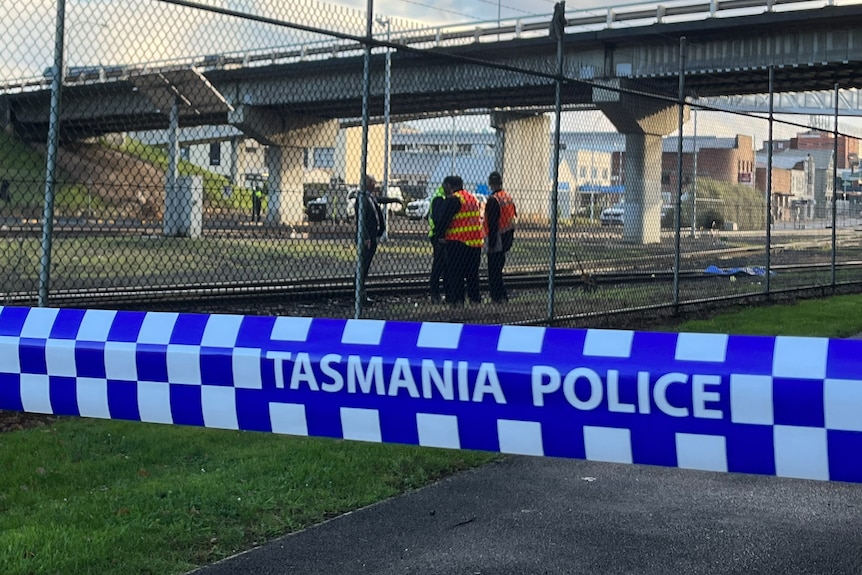 Blue and white police tape in front of a wire fence with men standing near railways lines in the background 