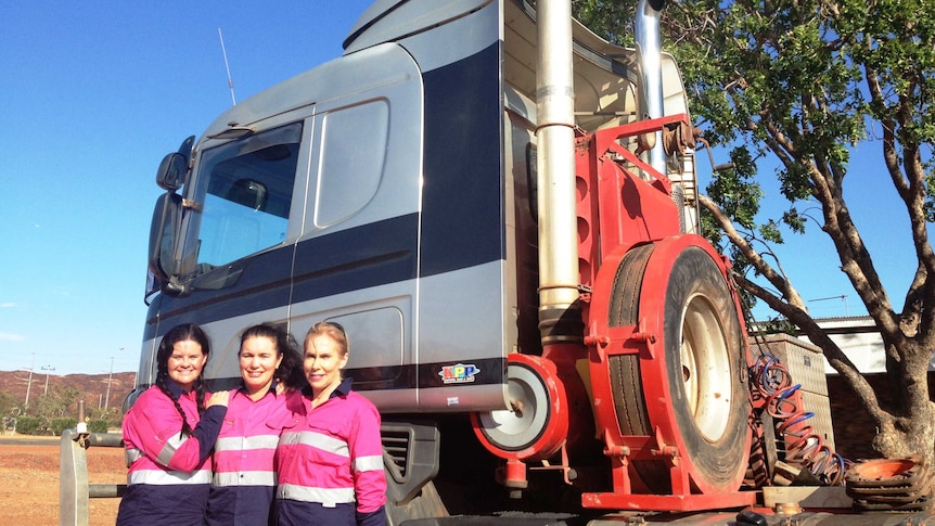 Three women wearing hot pink high-vis shirts stand in front of a truck cab