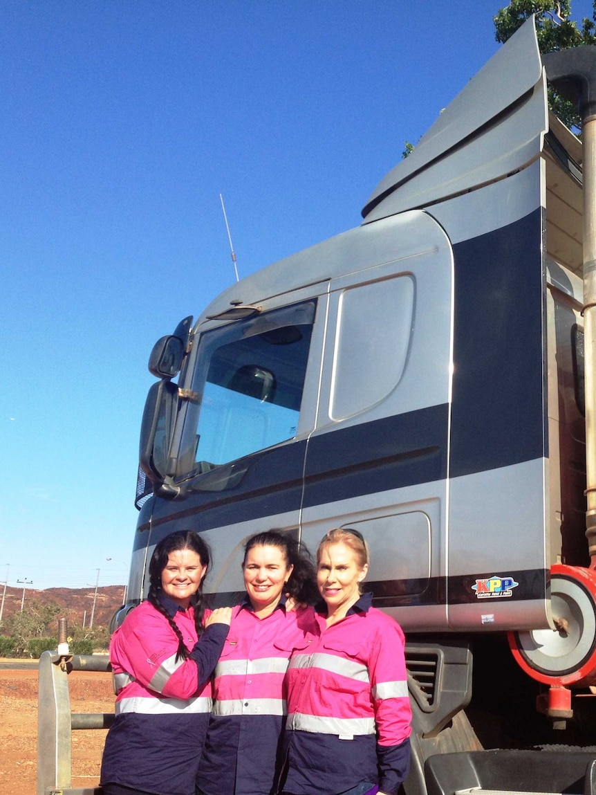 Three women wearing hot pink high-vis shirts stand in front of a truck cab