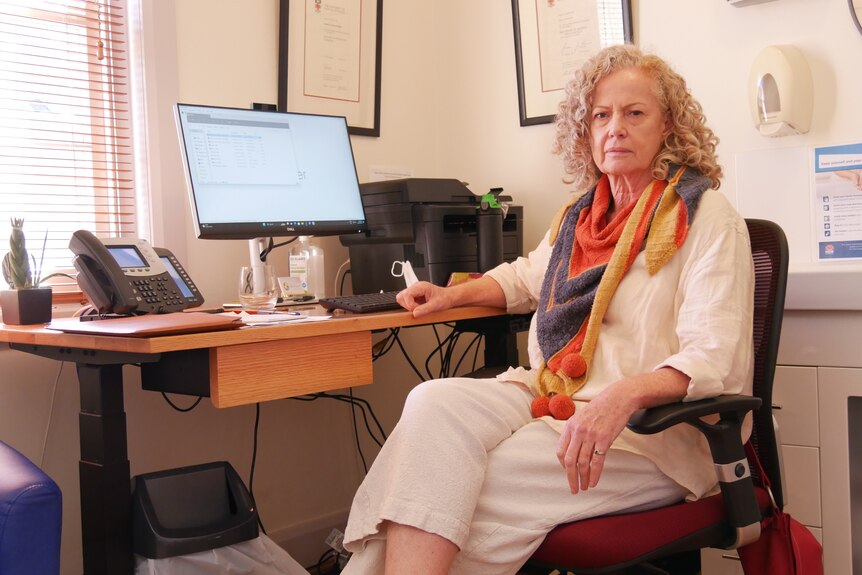 a woman sits at her desk in an office. She is wearing white linen clothing with a colourful scarf and curly hair