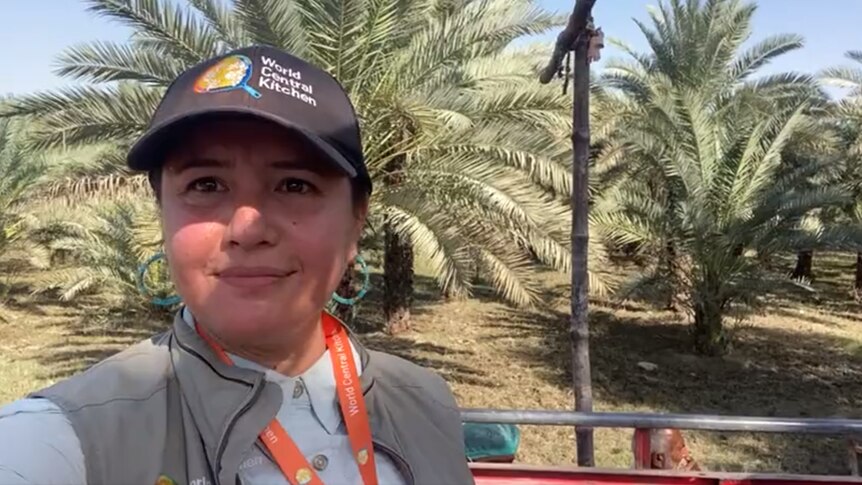 A woman wearing a World Central Kitchen cap points the camera at herself, palm trees in the background