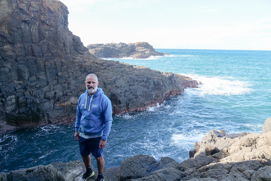 A man stands at the foot of a cliff, ocean behind him.