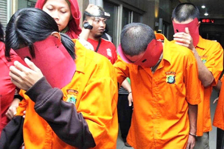 Three prisoners dressed in orange and wearing masks have their heads down as they walk.