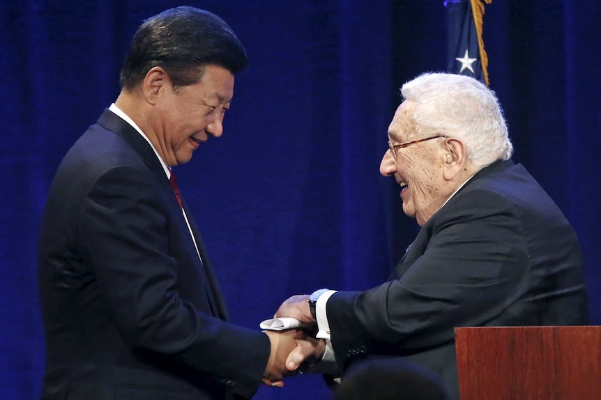 Two men shake hands while smiling, in front of a flag.