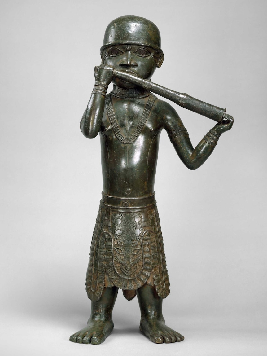 A brass sculpture of a figure holding a long horn to their mouth. The figure wears a skirt with a leopard pattern.