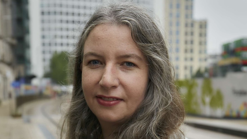 A white woman with shoulder length greying hair wearing a light jacket and black shirt against a background of large buildings