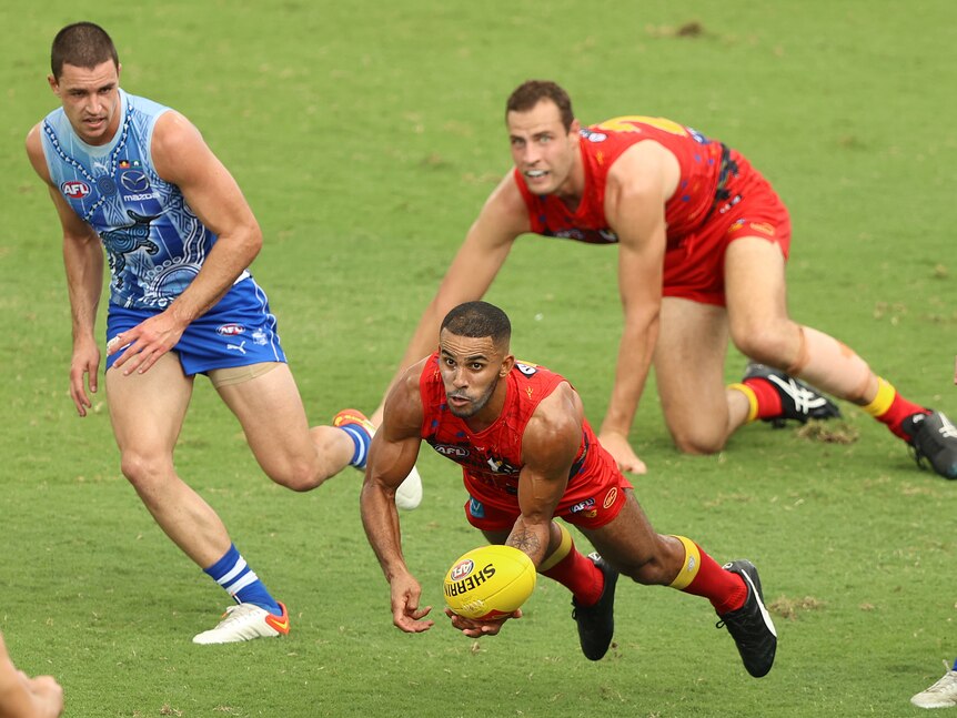 A Gold Coast AFL player launches himself forward off the ground getting ready to pump a handball away.